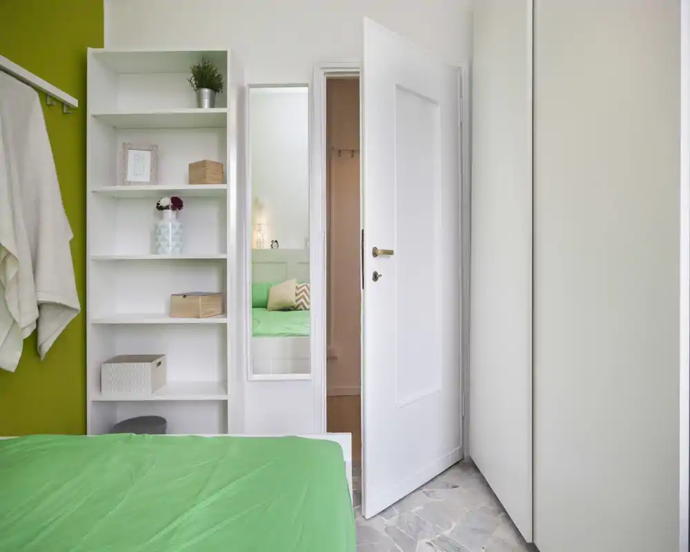 Dormitorio luminoso y ordenado con colcha verde, armario abierto y estanterías; la cama es el elemento principal y la habitación se siente fresca y acogedora.