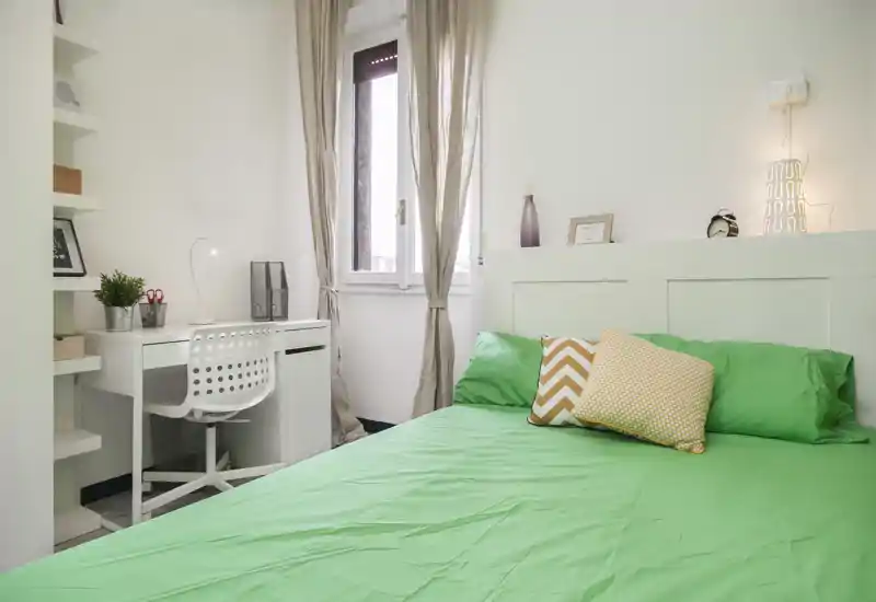 Dormitorio luminoso y ordenado con cama doble vestida de verde, cojines decorativos y un pequeño escritorio junto a la ventana — ideal para una estancia cómoda.