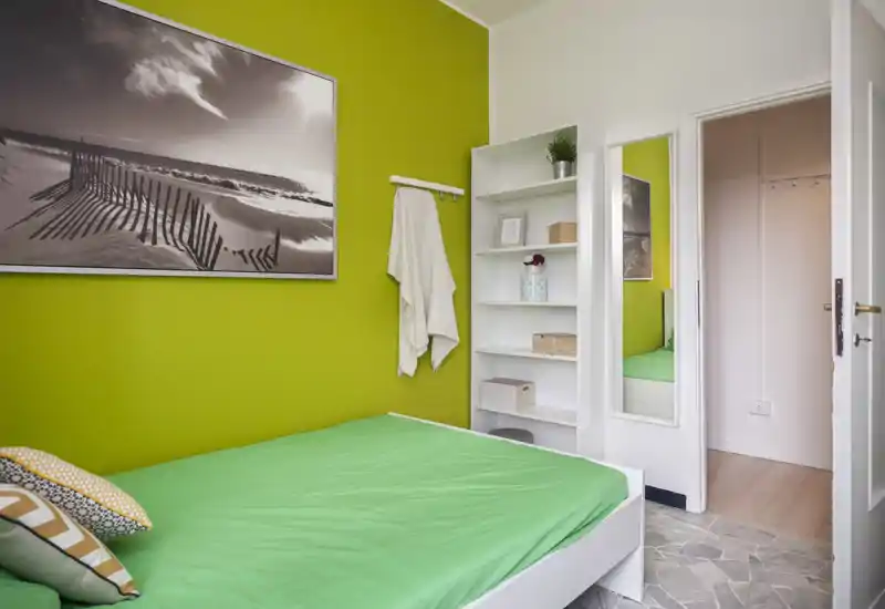 Dormitorio moderno y luminoso con pared verde, cama bien hecha con cojines de colores, estantería abierta y espejo de cuerpo entero junto a la puerta.