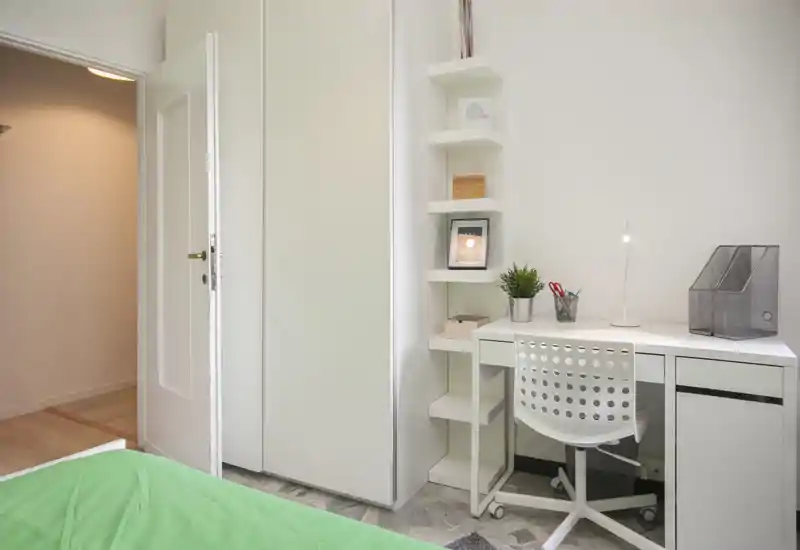 Rincón de dormitorio compacto con escritorio y silla blancos junto a un armario empotrado; se ve parcialmente una colcha verde en primer plano. Espacio luminoso, minimalista y ordenado, ideal para un estudiante o persona sola.