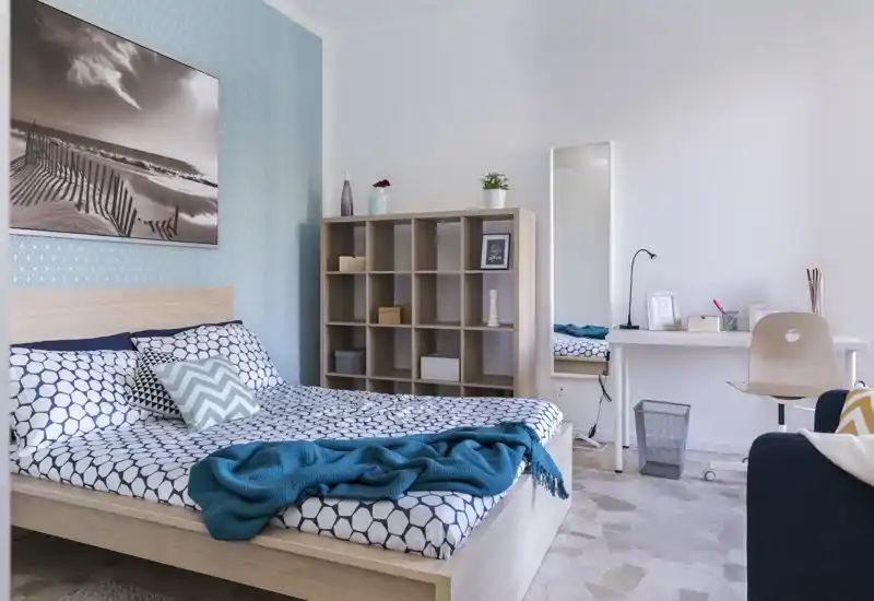 Camera da letto luminosa e curata con biancheria a fantasia, plaid turchese e struttura del letto in legno; libreria e angolo scrivania piccoli, puliti e accoglienti.