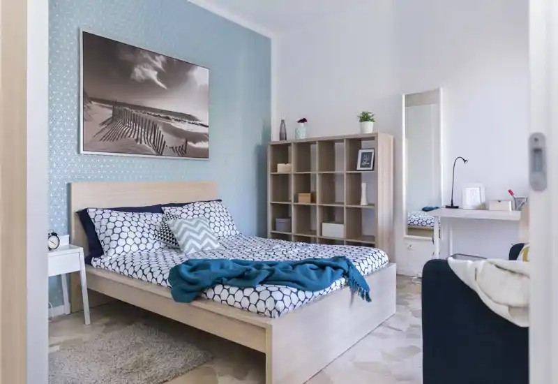Camera da letto luminosa e curata con parete decorativa, struttura letto in legno, tessili confortevoli e libreria — perfetta per annunci.