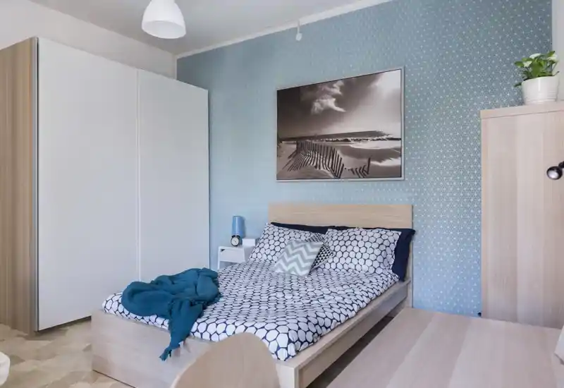 Camera da letto moderna e luminosa con carta da parati blu a motivi, letto matrimoniale ordinato con cuscini decorativi e coperta accogliente, oltre a ampio spazio contenitivo (guardaroba e mobile). Linee pulite e colori armoniosi creano un’atmosfera rilassante perfetta per annunci.