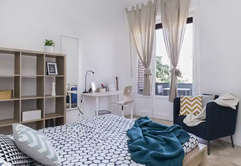 Camera da letto luminosa e ben arredata con letto matrimoniale a motivi, plaid e cuscini accoglienti; una scrivania e una poltrona vicino a una grande finestra/porta che dà sul balcone con luce naturale.