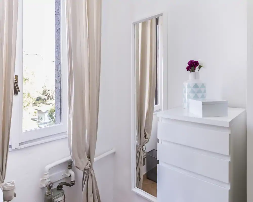 Angolo di camera da letto luminoso con comò bianco, specchio a figura intera e tende beige che lasciano entrare la luce naturale; arredo minimal con un vaso decorativo per un tocco accogliente.