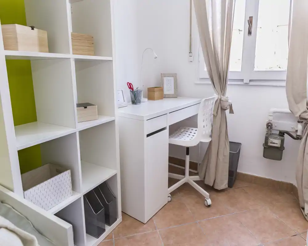 Angolo lavoro compatto e luminoso con scrivania bianca, sedia ergonomica e scaffalatura — ideale per un piccolo ufficio domestico o angolo studio con luce naturale.