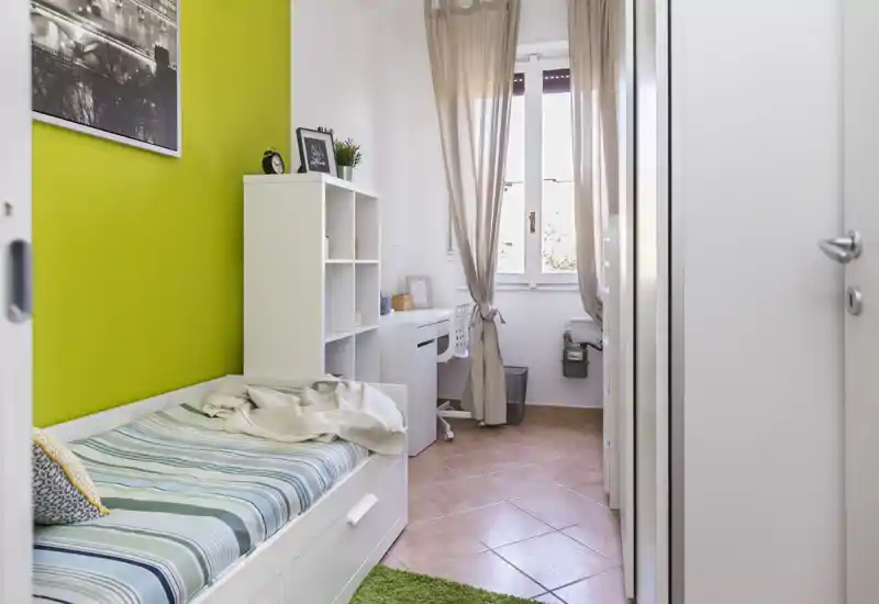 Chambre compacte et lumineuse avec un lit simple contre un mur vert, étagères organisées et un petit bureau près de la fenêtre apportant de la lumière naturelle.