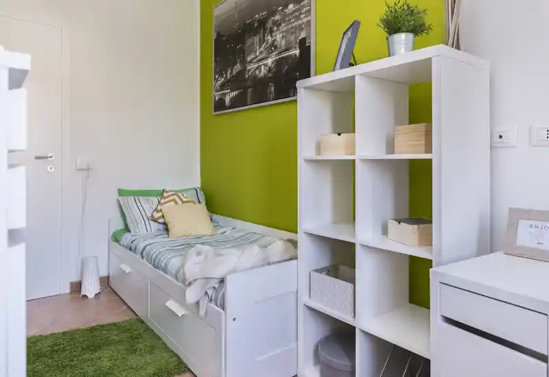 Chambre simple compacte avec un lit blanc, un mur vert accrocheur et des étagères bien rangées. L’espace est lumineux et soigné, avec des coussins et textiles confortables pour un séjour douillet.