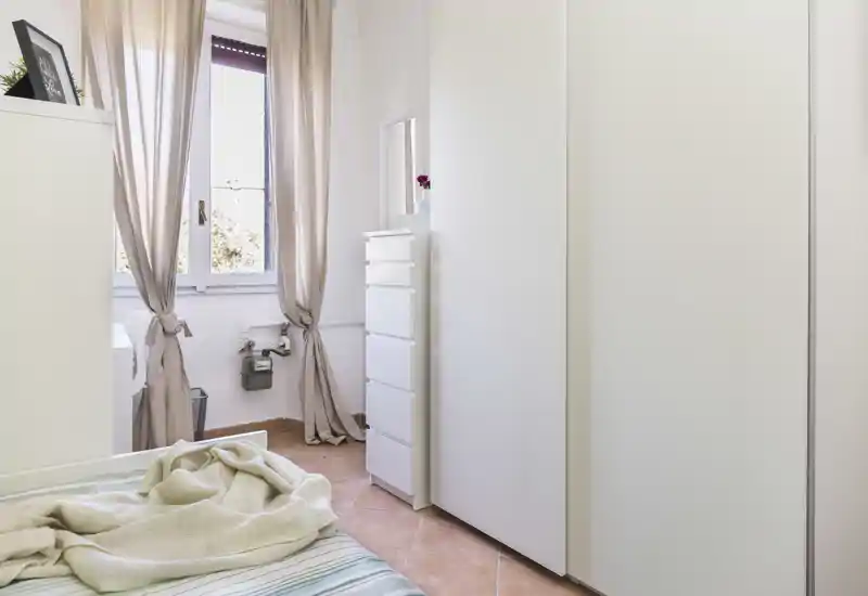 Coin de chambre lumineux et compact avec textiles neutres, lit partiellement visible et grands rangements blancs près d'une fenêtre ensoleillée.