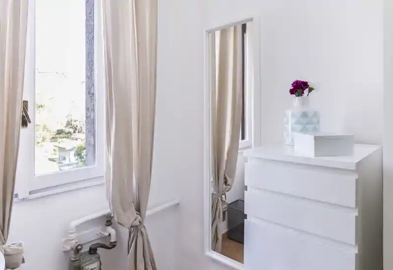 Coin de chambre lumineux avec une commode blanche, un miroir pleine longueur et des rideaux beige laissant entrer la lumière naturelle ; décoration minimaliste avec un vase décoratif pour une touche chaleureuse.