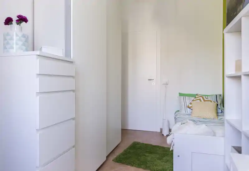 Petite chambre lumineuse avec un lit simple garni de coussins à motifs et d'un plaid cosy, des rangements blancs et un tapis vert pour un style frais et minimaliste.