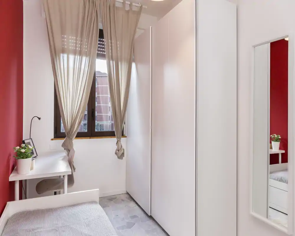 Camera piccola e ben arredata con letto singolo, grande armadio bianco, scrivania vicino alla finestra e luce naturale calda — ideale per studenti o soggiorni brevi.