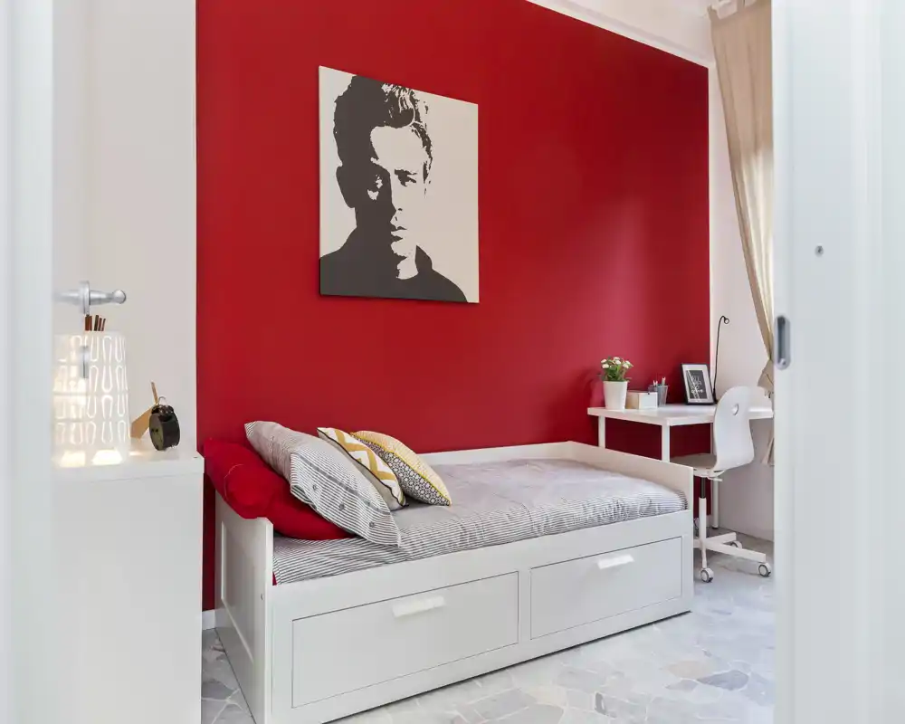 Camera singola luminosa e moderna con letto a giorno bianco, parete rossa vivace e una piccola scrivania vicino alla finestra — ben illuminata e arredata con gusto.