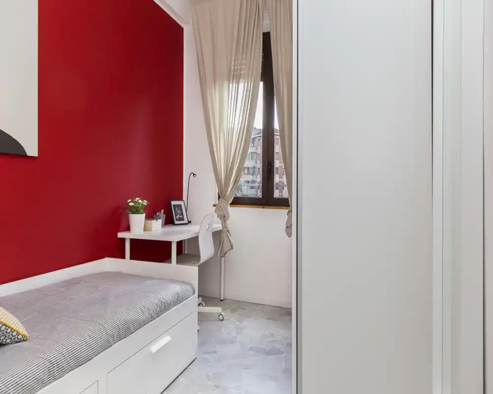 Camera singola compatta con letto, cassetto contenitore bianco sotto il letto, piccolo scrittoio vicino alla finestra e parete d'accento rossa; luce naturale abbondante e arredo curato.