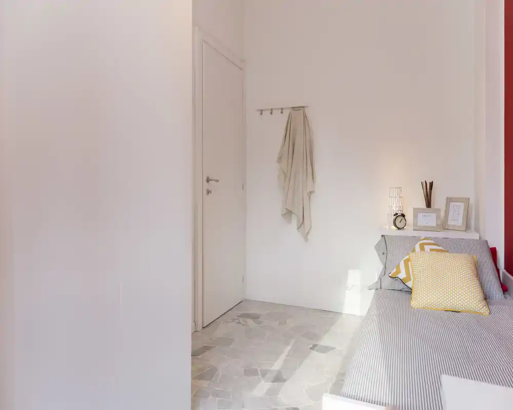 Camera da letto compatta e luminosa con letto singolo in lenzuola a righe e cuscini gialli, mensola laterale con oggetti decorativi e luce naturale.