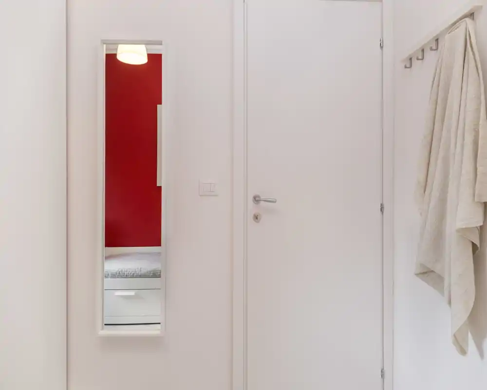 Scatto interno luminoso e minimal con una porta bianca e uno specchio stretto a parete che riflette una parte del letto con la parete rossa sullo sfondo.