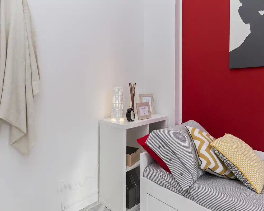 Angolo camera luminoso e curato con cuscini decorativi, mensola come comodino, lampada e parete rossa a contrasto — perfetto per una singola accogliente o una stanza per gli ospiti.
