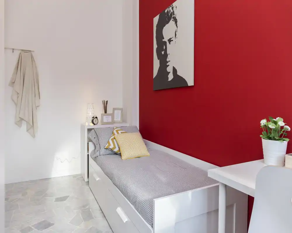 Camera singola luminosa e curata con letto bianco, cuscini decorativi e una splendida parete rossa; dispone di scrivania e comodino, perfetta per un appartamento compatto.