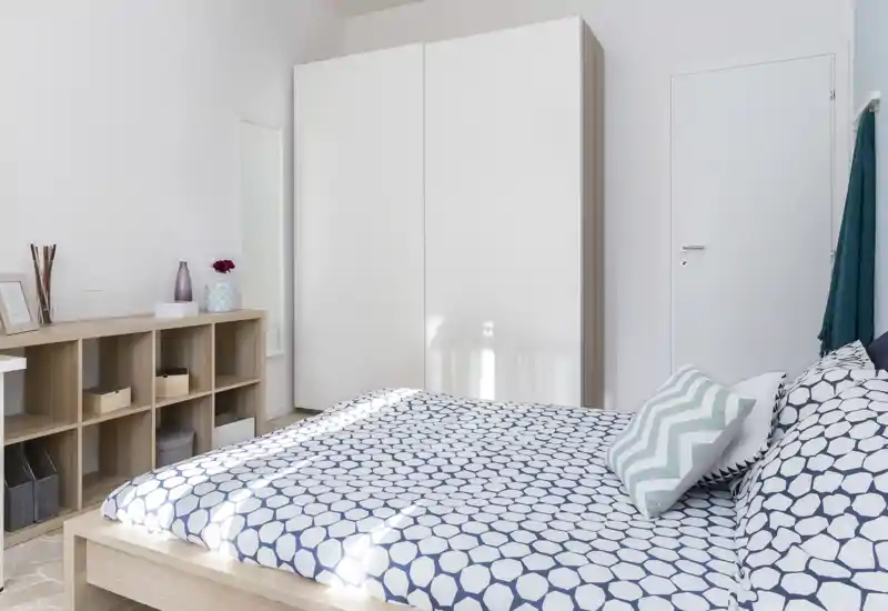 Camera da letto luminosa e moderna con copriletto a motivo, cuscini decorativi e un ampio armadio bianco; scaffalatura minimale per riporre oggetti.