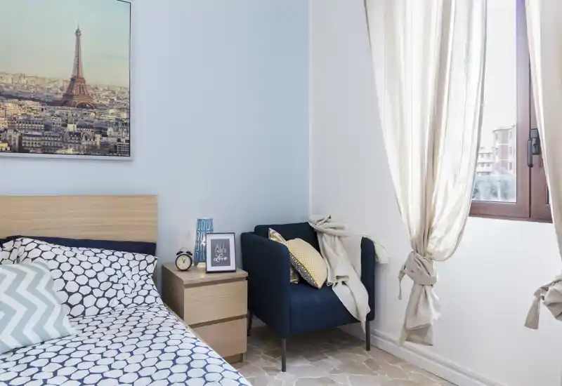 Bright, neatly styled bedroom with a patterned duvet, wooden headboard and a cozy armchair by the window letting in natural light.