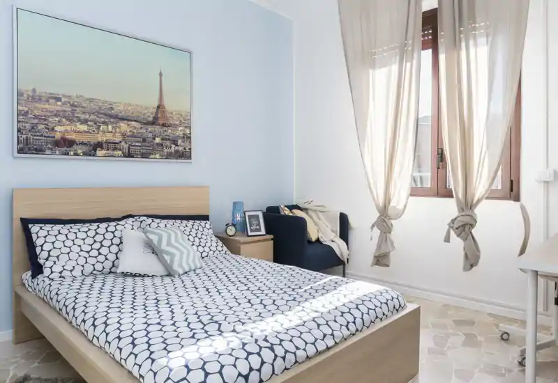 Camera da letto luminosa e curata con struttura del letto in legno, biancheria a fantasia, comodino e poltrona vicino alla finestra. Tende leggere e ampia finestra garantiscono molta luce naturale, creando un ambiente arioso e accogliente perfetto per annunci.