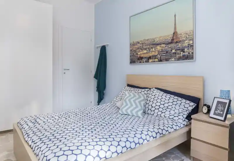 Camera da letto luminosa e di design con struttura del letto in legno, biancheria a motivo e un quadro decorativo. L'ambiente è fresco e accogliente, perfetto per un annuncio di affitto.
