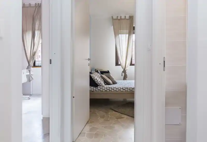 Bright, cozy bedroom seen through an interior doorway: double bed with patterned bedding, soft pillows and natural light from a curtained window.