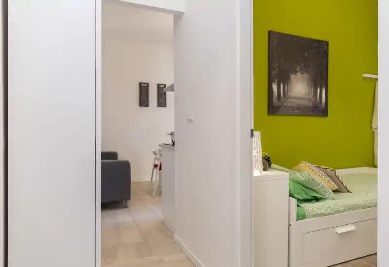 Compact, well-styled bedroom corner with a green accent wall, a tidy single bed with layered cushions and a small bedside surface — ideal for a cozy single occupancy.