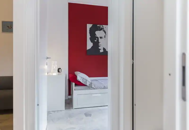 Bright single bedroom seen through a doorway with a red accent wall, a neatly made bed with pillows and a small white nightstand — clean, modern and inviting.