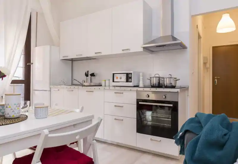 Modern compact kitchen with white cabinets, oven and microwave. A small dining table with chairs and soft textiles creates a welcoming, well-lit space ideal for short-term rentals.