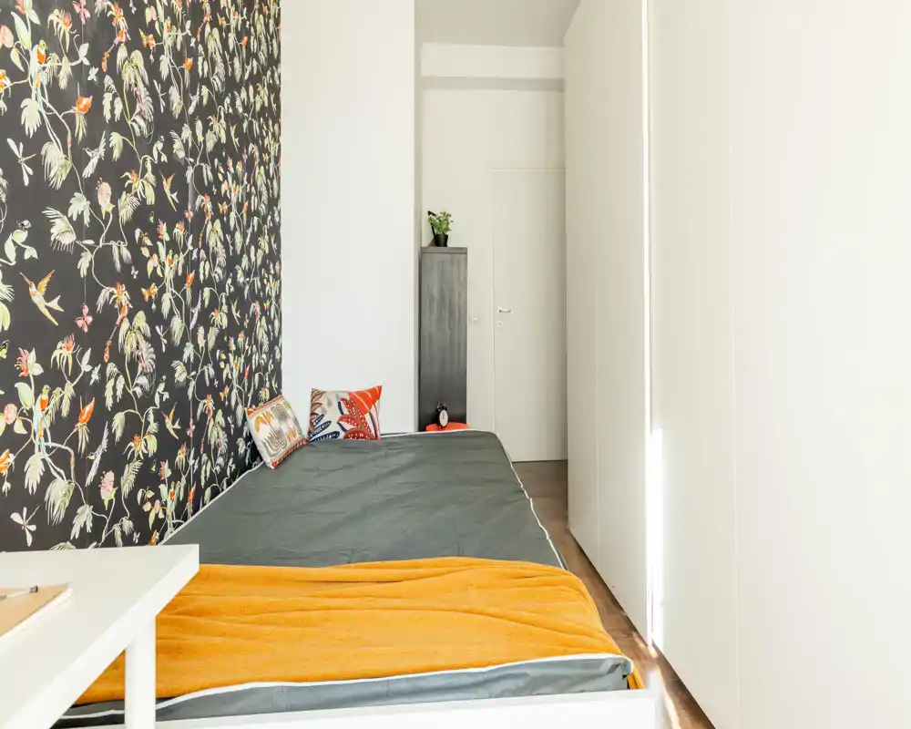 Dormitorio moderno y luminoso con cama individual junto a una pared floral, cojines coloridos y una cálida manta naranja. Distribución minimalista ideal para una persona.