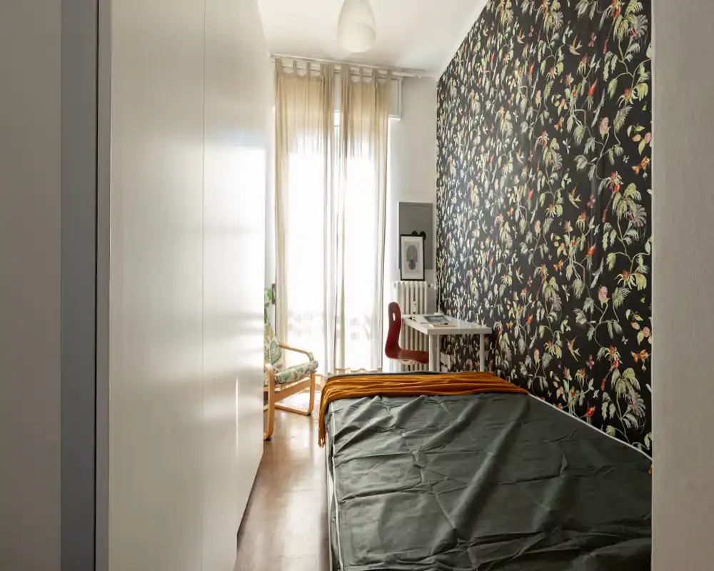 Dormitorio estrecho y luminoso con papel pintado decorativo, una cama individual y un pequeño escritorio junto a la ventana. La luz natural filtrada por las cortinas aporta una atmósfera tranquila y con estilo, adecuada para una persona o alquiler temporal.