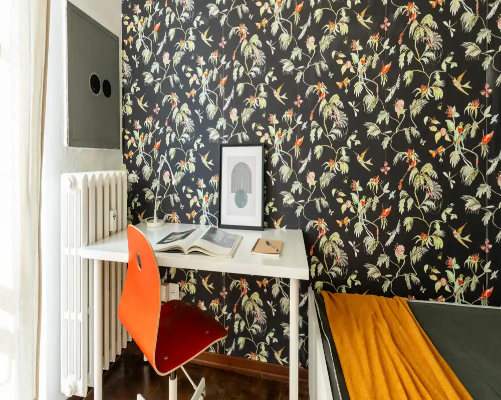 Rincón de dormitorio compacto con un escritorio blanco, silla naranja y cama parcialmente visible con una manta mostaza; papel pintado floral que añade calidez y estilo.