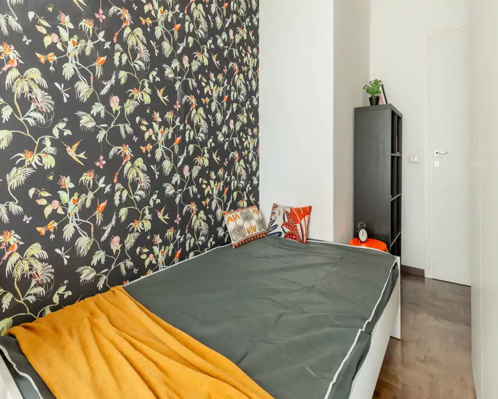 Dormitorio compacto y con estilo con pared decorativa estampada, cama individual con ropa de cama oscura y manta mostaza cálida; estantería ordenada y cojines decorativos que completan un ambiente acogedor.