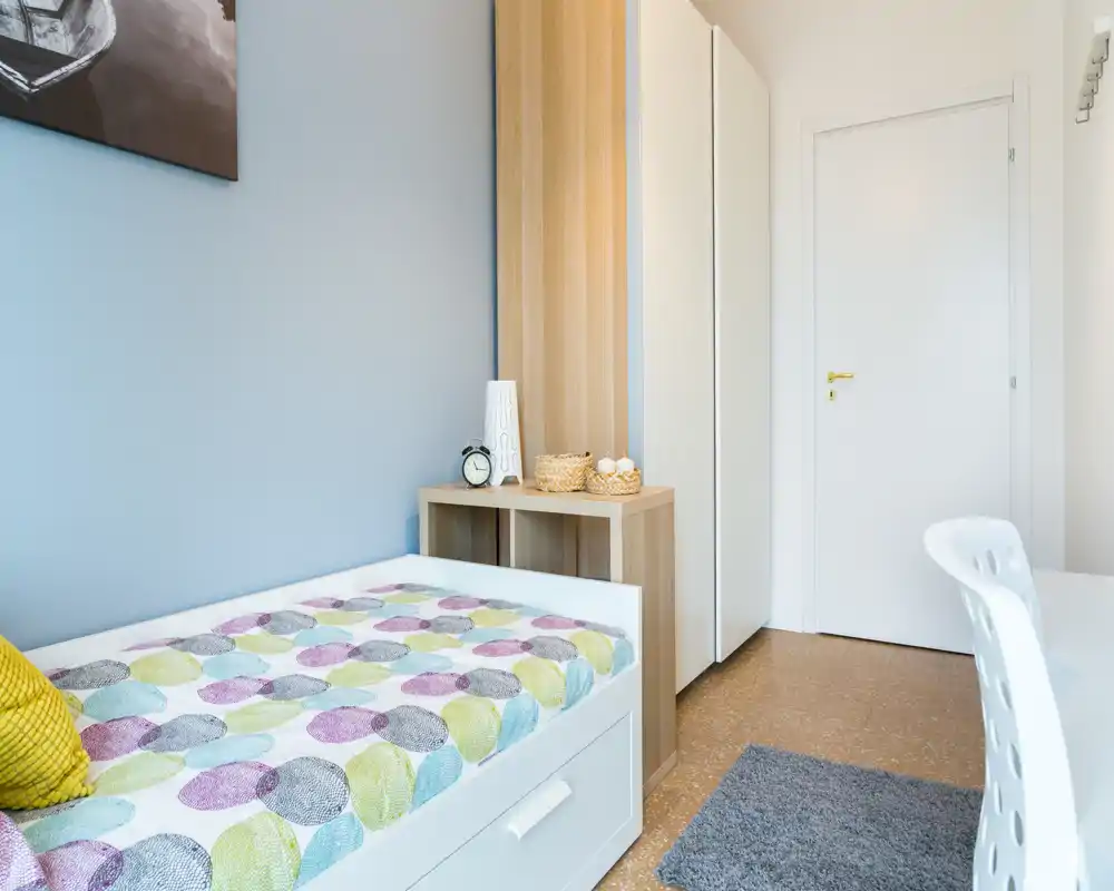 Habitación individual luminosa y ordenada con colcha de estampado, estantería con objetos decorativos y una zona de escritorio minimalista — perfecta para un alquiler acogedor y compacto.