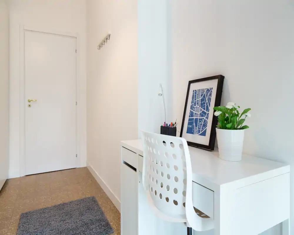 Entrada luminosa y minimalista con un pequeño escritorio y silla, percheros junto a la puerta y felpudo — estilo moderno y limpio, ideal para un apartamento compacto.