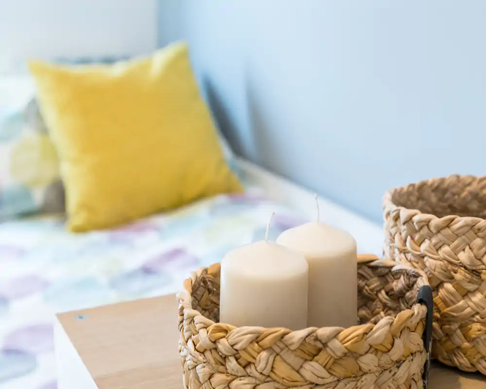 Detalle de dormitorio acogedor con cestas trenzadas y velas decorativas en primer plano y una cama con cojines de colores desenfocada al fondo.