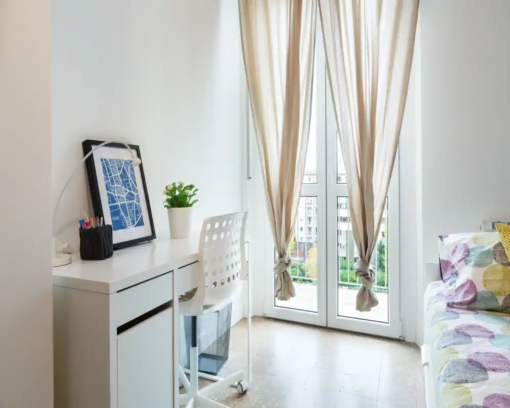 Rincón de dormitorio luminoso y aireado con escritorio y silla junto a puertas hasta el suelo; vista parcial de la cama con cojines coloridos y ambiente moderno y acogedor.