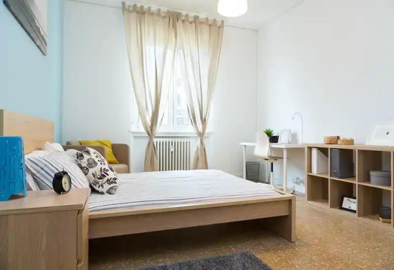 Camera da letto luminosa e ben allestita con letto in legno, comodino e un piccolo angolo studio vicino alla finestra. Toni neutri e cuscini decorativi creano un'atmosfera calma e accogliente.