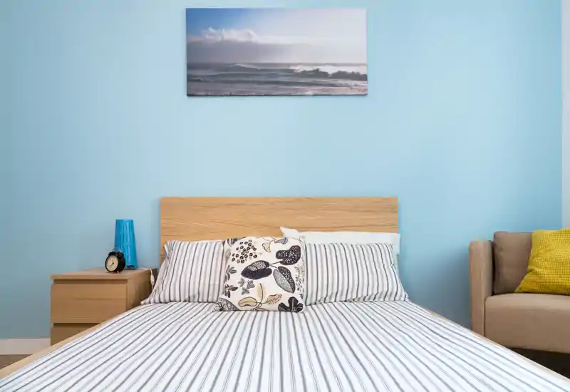 Camera da letto luminosa e ben arredata con un letto matrimoniale ordinato, lenzuola a righe, comodino con lampada e una stampa marina sopra la testata — perfetta per un soggiorno rilassante.