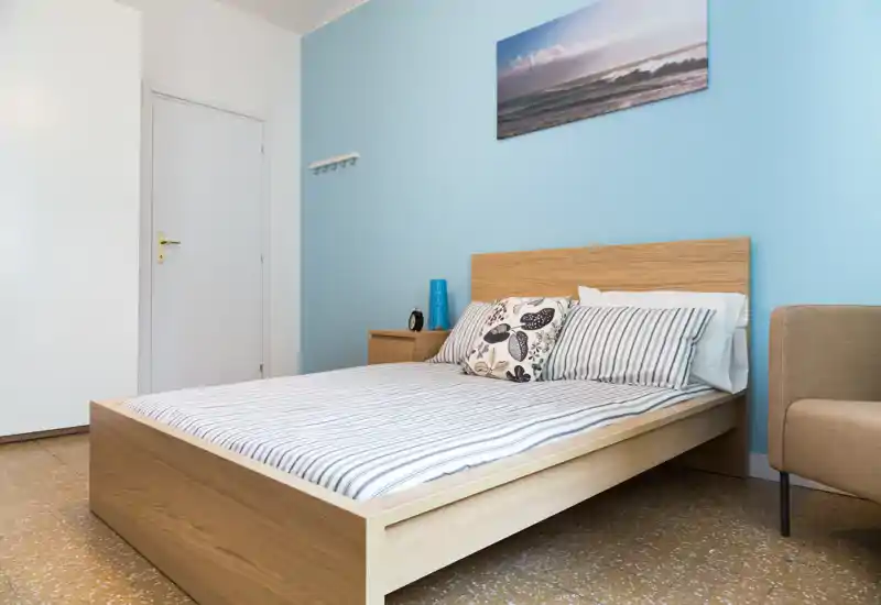 Camera da letto luminosa e ordinata con letto matrimoniale in legno, biancheria a righe e cuscini decorativi su una parete azzurra; comodino e lampada completano l’ambiente accogliente.