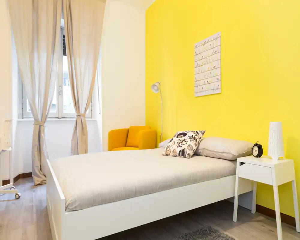 Camera da letto luminosa e ordinata con letto bianco, comodino e una parete gialla vivace. La luce naturale dalla finestra e una poltroncina accogliente creano un'atmosfera ideale per annunci di affitto.
