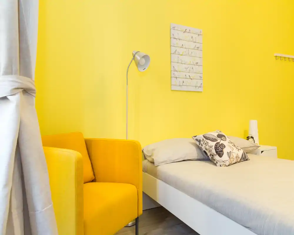 Camera da letto luminosa e moderna con letto singolo in lino neutro e una poltrona gialla vivace. Pareti gialle calde, comodino con lampada e cuscino decorativo per un ambiente accogliente.