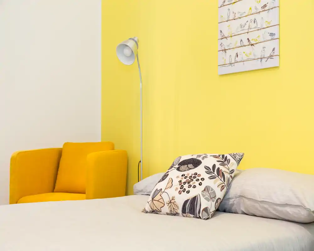 Angolo camera luminoso e accogliente con letto ben rifatto, cuscino decorativo a motivo, poltrona gialla e lampada da terra su una parete gialla vivace.