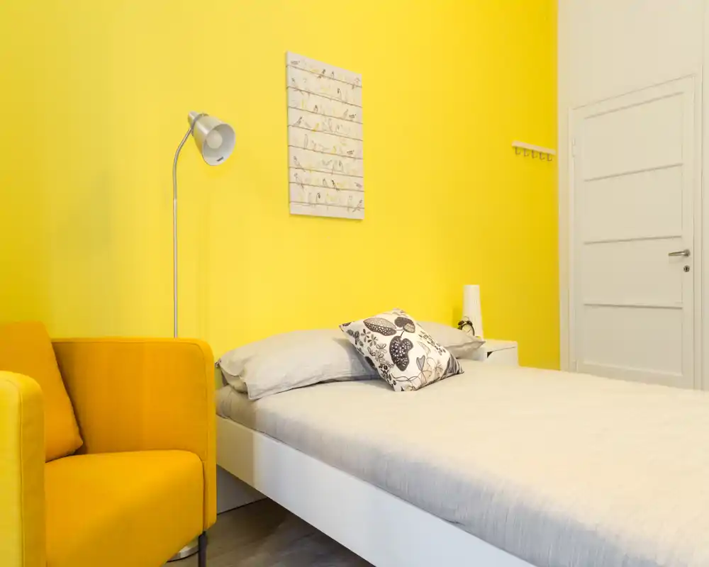 Camera da letto singola moderna e luminosa con letto bianco, cuscino decorativo e parete accentuata in giallo; poltrona gialla e comodino per un ambiente accogliente.