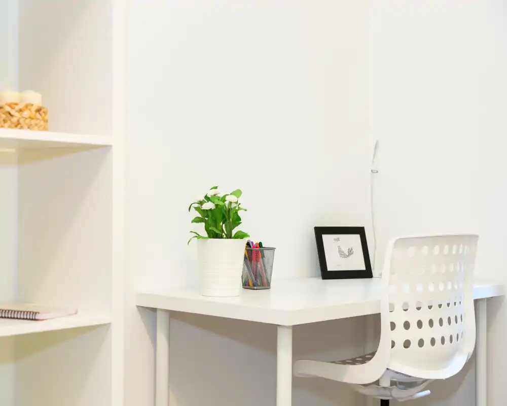 Angolo studio luminoso e minimal con scrivania bianca, sedia traforata, una pianta in vaso e una piccola stampa incorniciata: spazio di lavoro moderno e ordinato.