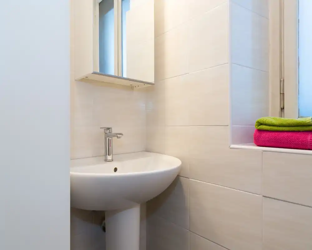 Rincón de baño moderno y compacto con lavabo de pedestal, armario con espejo y toallas coloridas dobladas en el alféizar — luminoso, limpio y funcional.