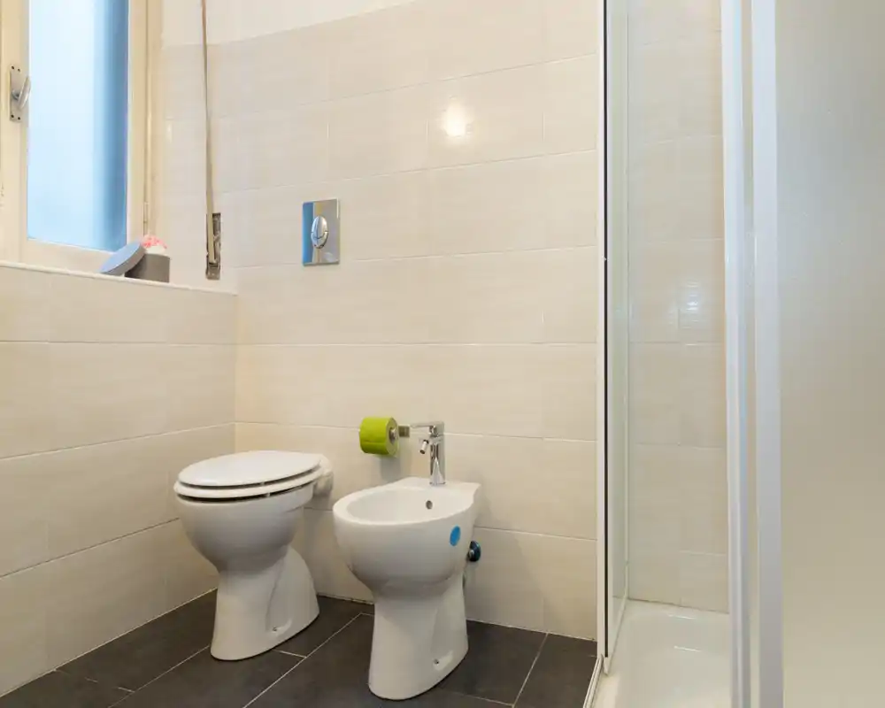 Esquina de baño moderna y limpia con inodoro, bidé y mampara de ducha de vidrio, azulejos neutros y suelo oscuro.