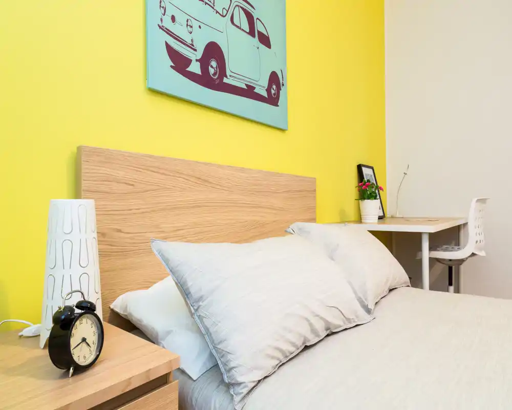 Dormitorio luminoso y bien decorado con cabecero de madera clara, ropa de cama neutra y una pared amarilla; mesita con despertador y un pequeño escritorio en la esquina.