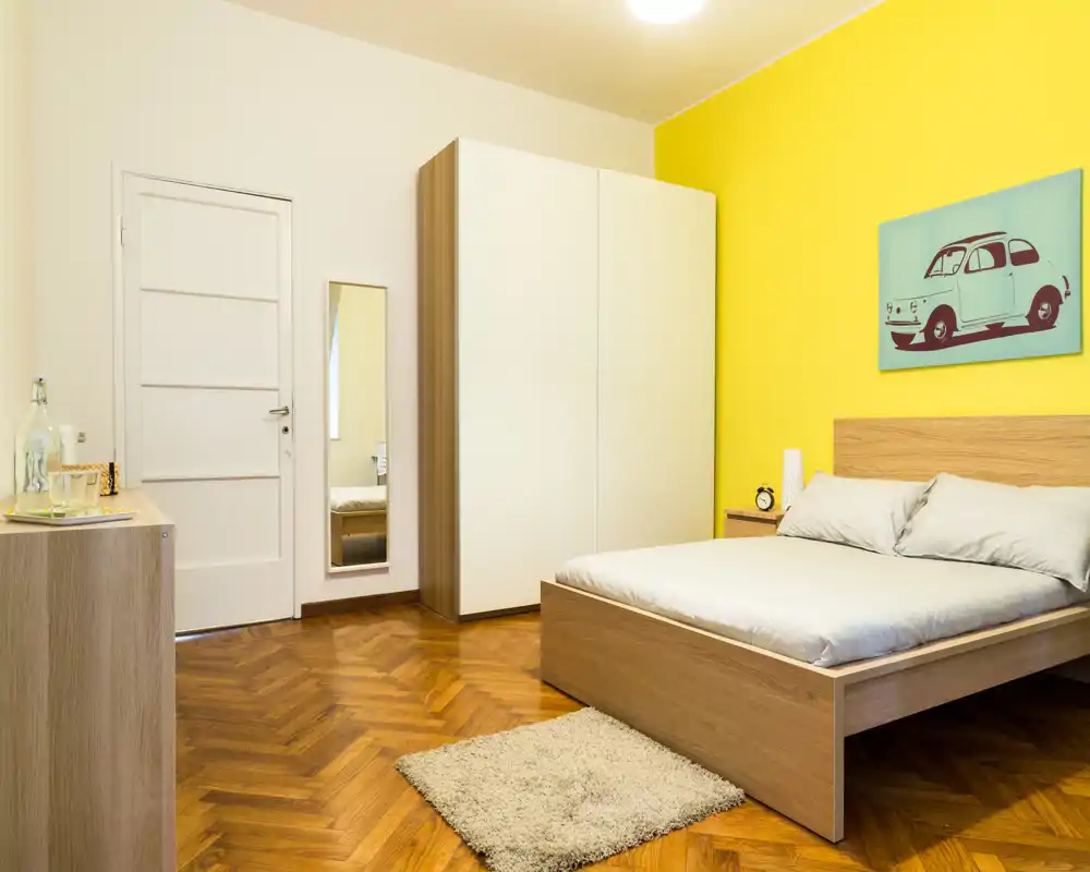 Dormitorio luminoso y bien decorado con cama doble de madera, mesita y armario frente a una pared amarilla. El suelo de parquet y la pequeña alfombra aportan calidez y un ambiente moderno y acogedor.